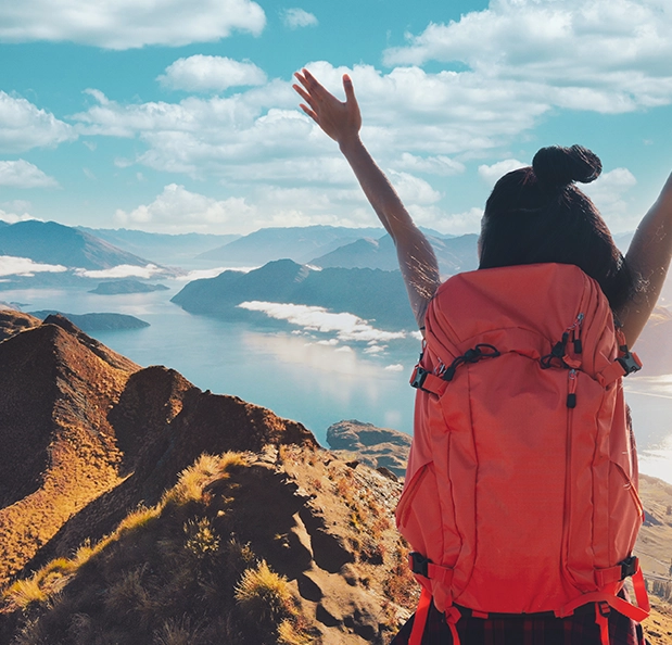 A woman wearing a bright orange pack stands atop a tussocky ridge-line, arms in the air